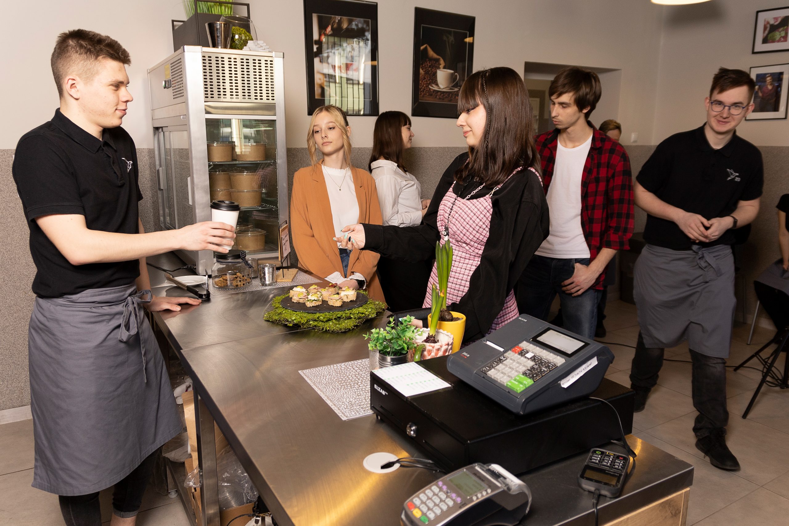 bistro, młodzi ludzie stoją przy ladzie, reszta stoi za nimi, kelner podaje kawę w papierowym kubku