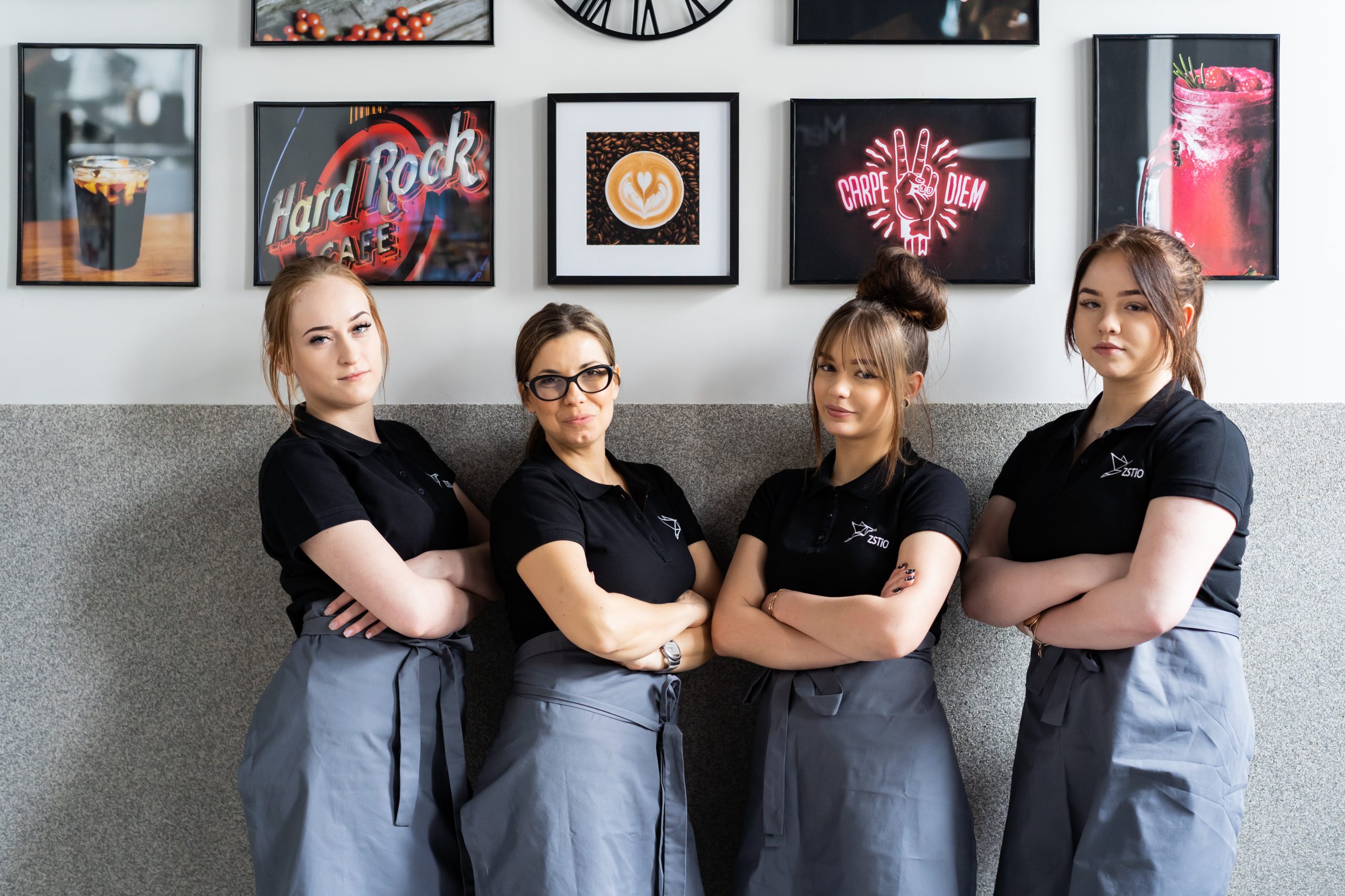 cztery kobiety ubrane w czarne koszulki z logo szkoły, na drugim planie obrazki na ścianie