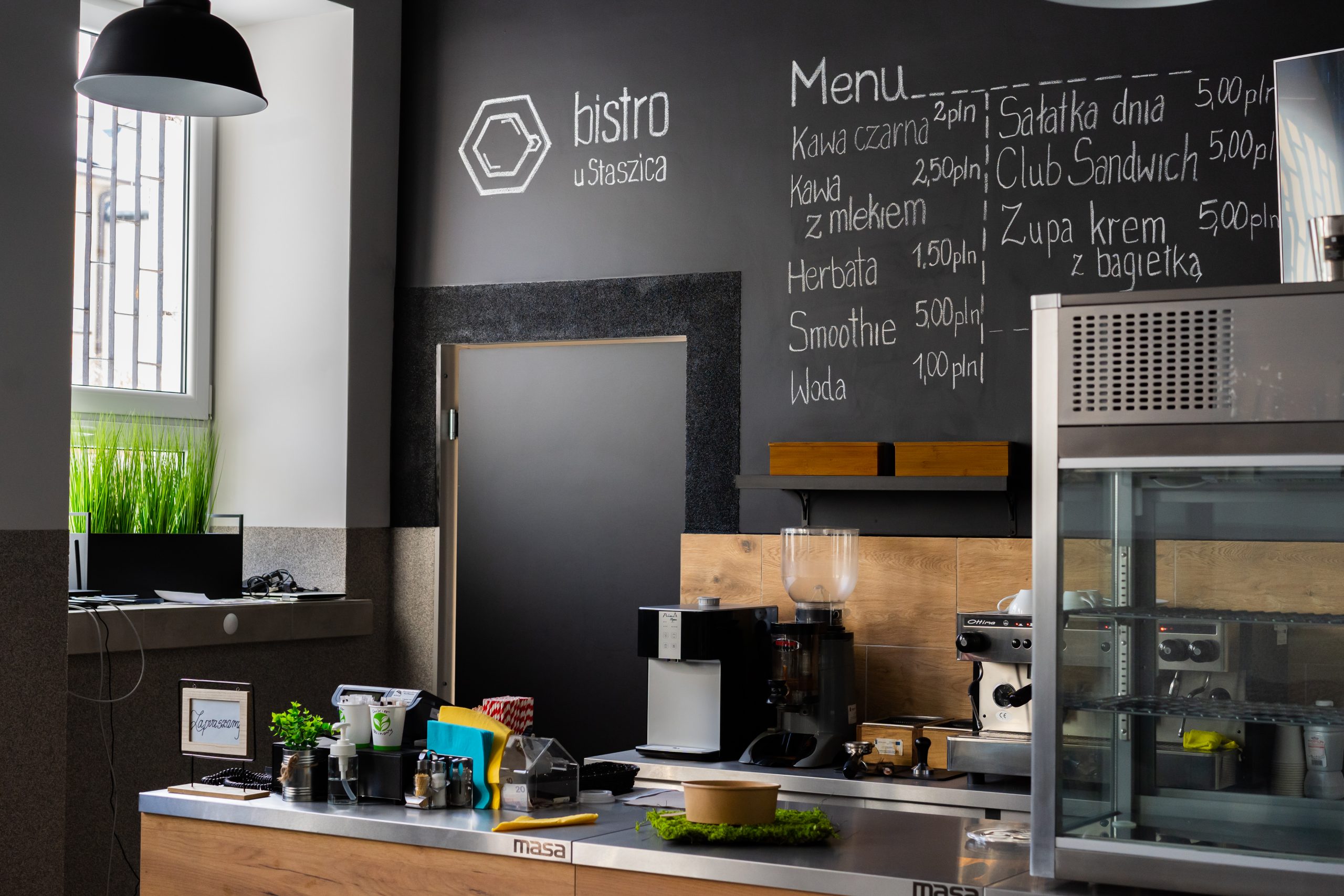 wnętrze bistro, czarna lampa, sztuczne kiaty w czarnej donicy na parapecie, lodówka, lada