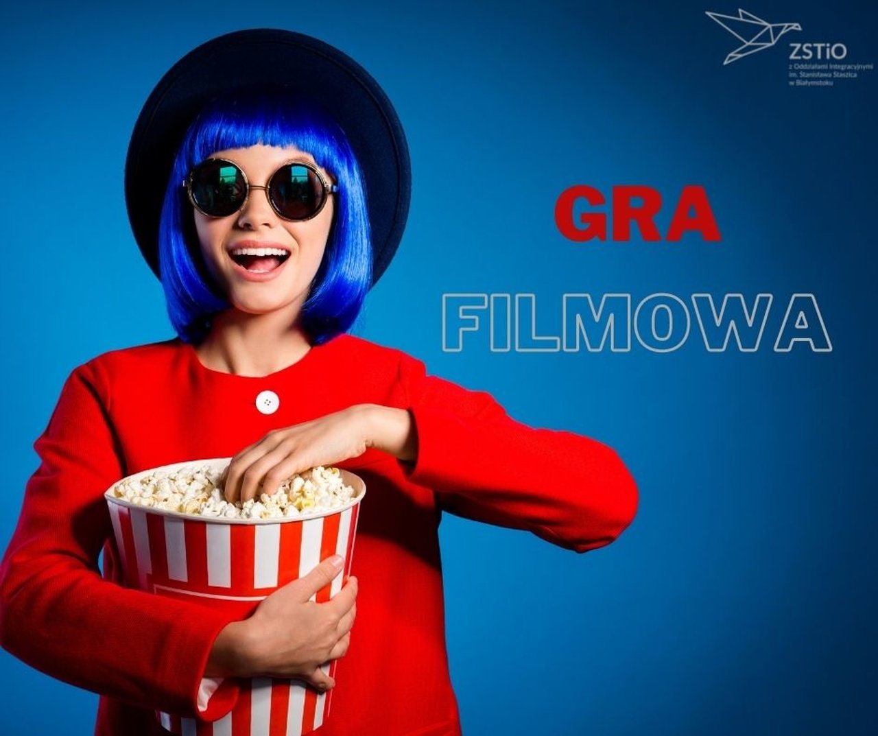 niebieskie tło, na pierwszym planie dziewczyna w kapeluszu z niebieskimi włosami, w czerwonej koszuli trzyma popcorn