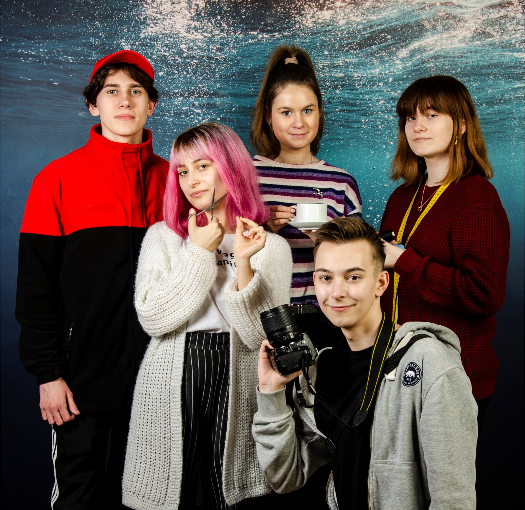 5 osób, chłopak w niebiesko-czerwnonej bluzie z czerwoną czapką z daszkiem, dziewczyna w różowych włosach trzyma w ręku nożyczki, chłopak w szarej bluzie trzyma aparat foto, dziewczuna w kucyku trzyma filiżankę