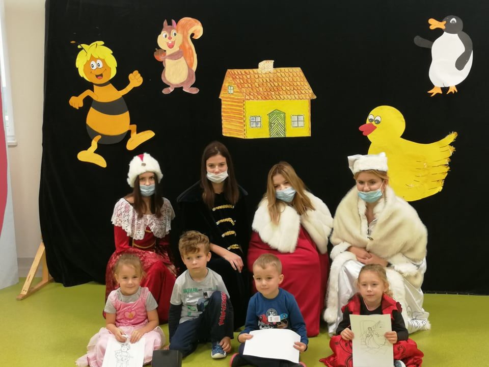 w tle zwierzęta, na pierwdszym planie siedzą dzieci, z tyłu cztery dziewczyny przebrane w historyczne stroje