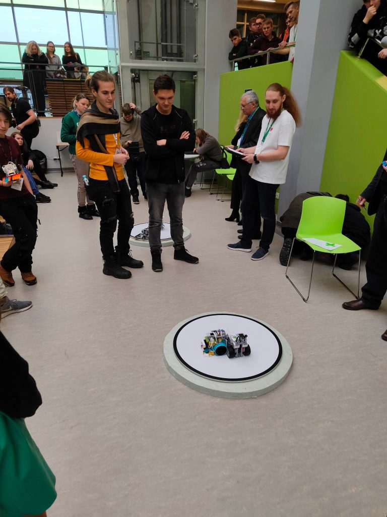 grupa ludzi, mężczyźni stoją, w rękach trzymają piloty, na ziemi roboty z klocków lego