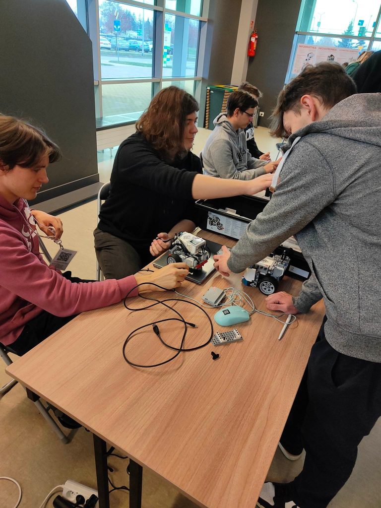 grupa ludzi, mężczyźni stoją, w rękach trzymają piloty, na stole roboty z klocków lego