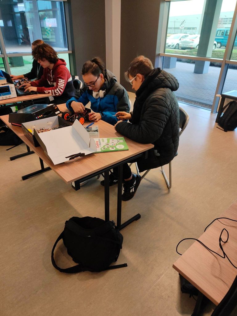 grupa ludzi, mężczyźni stoją, w rękach trzymają piloty, na stole roboty z klocków lego