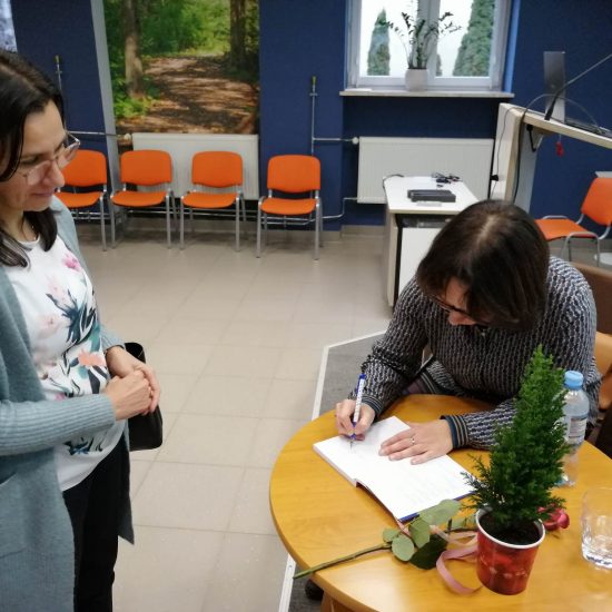 kobieta siedzi przy stoliku i podpisuje książę, druga kobieta stoi obok