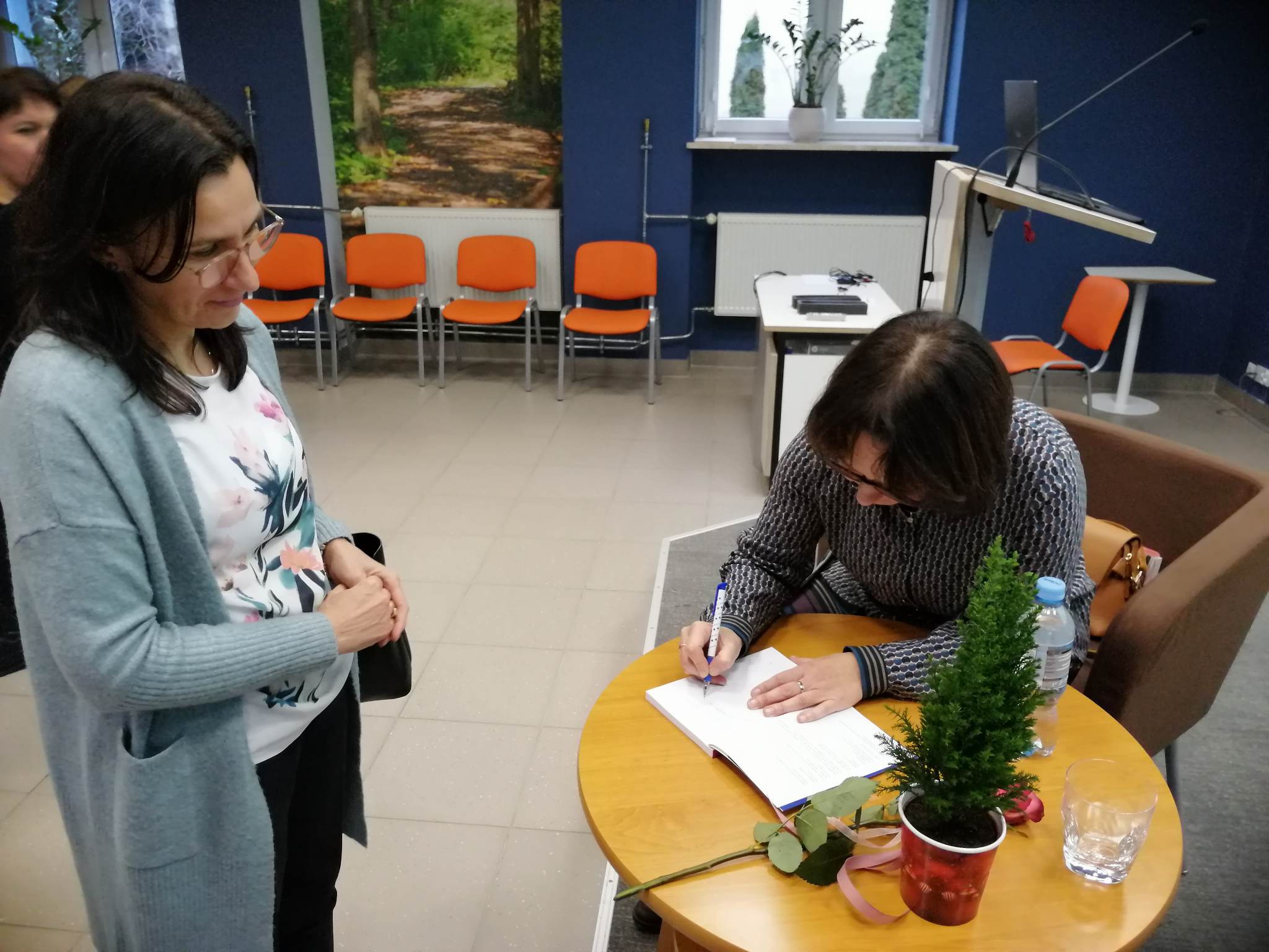 kobieta siedzi przy stoliku i podpisuje książę, druga kobieta stoi obok