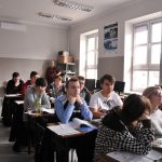 uczniowie siedzący w ławkach, w tle jasne okna