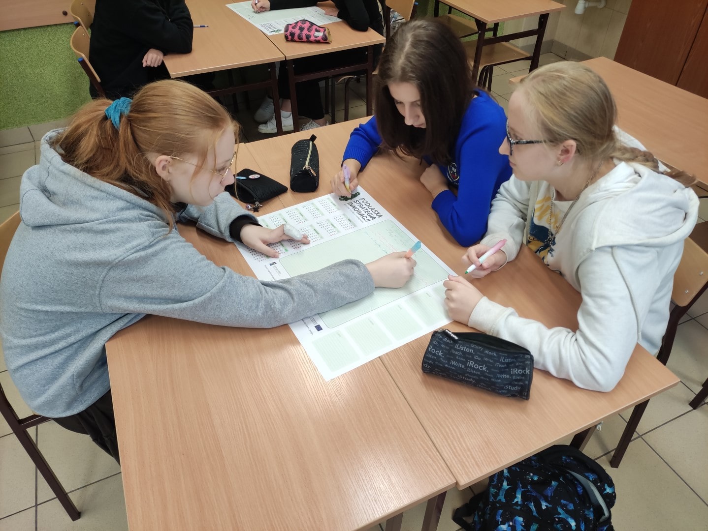 trzy dziewczyny siedzxą przy łąwce, na ławce karta pracy, piórniki