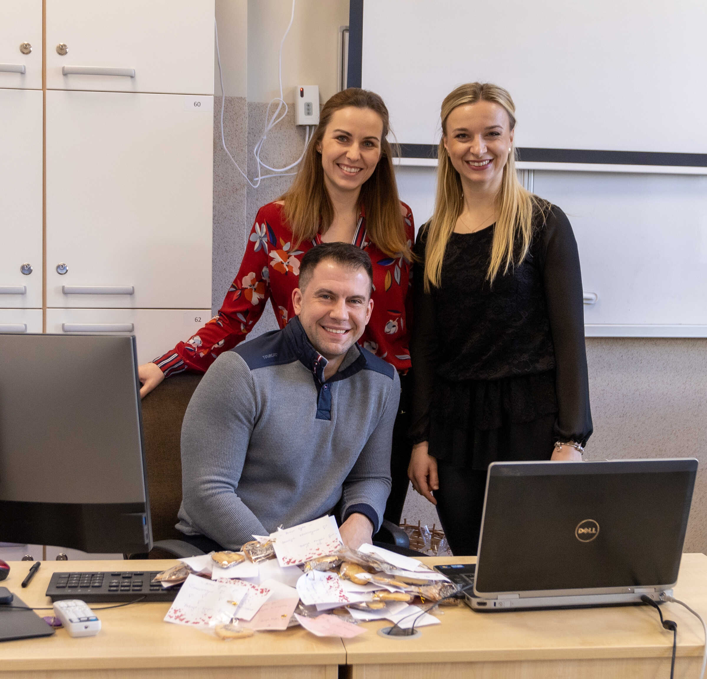 mężczyzna siedzi przy biurku, za nim stoją dwie młode kobiety, na stole ciastka, komputery