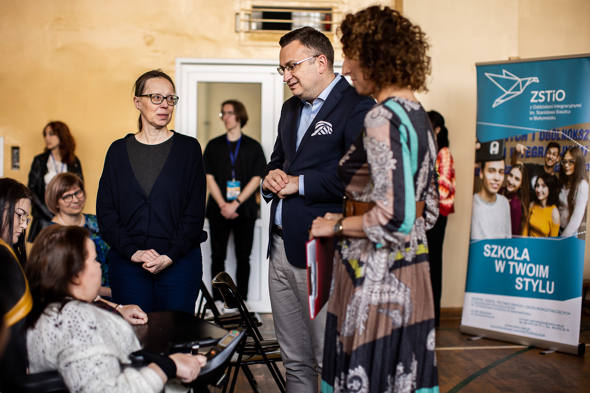 grupa osób, część siedzi na krzesłąch, część stoi na sali gimnastycznej