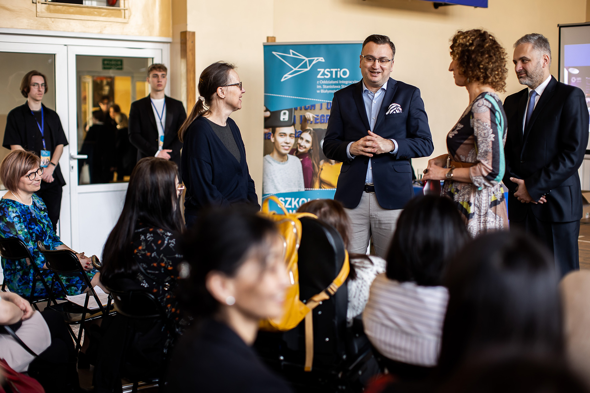 grupa osób, część siedzi na krzesłąch, część stoi na sali gimnastycznej