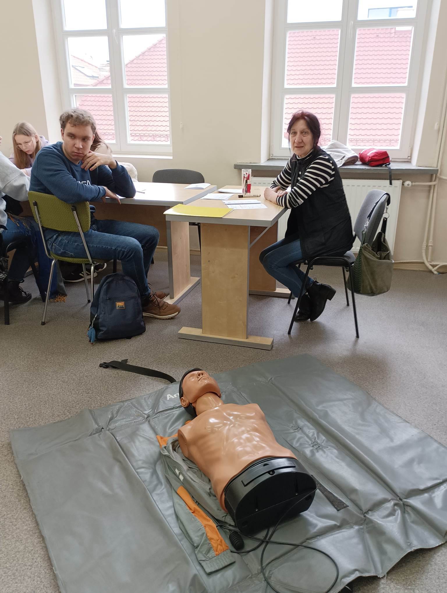 na pierwszym planie manekim, na drugim kobieta siedzi przy biurku, z lewej młodziez siedzi przy ławkach