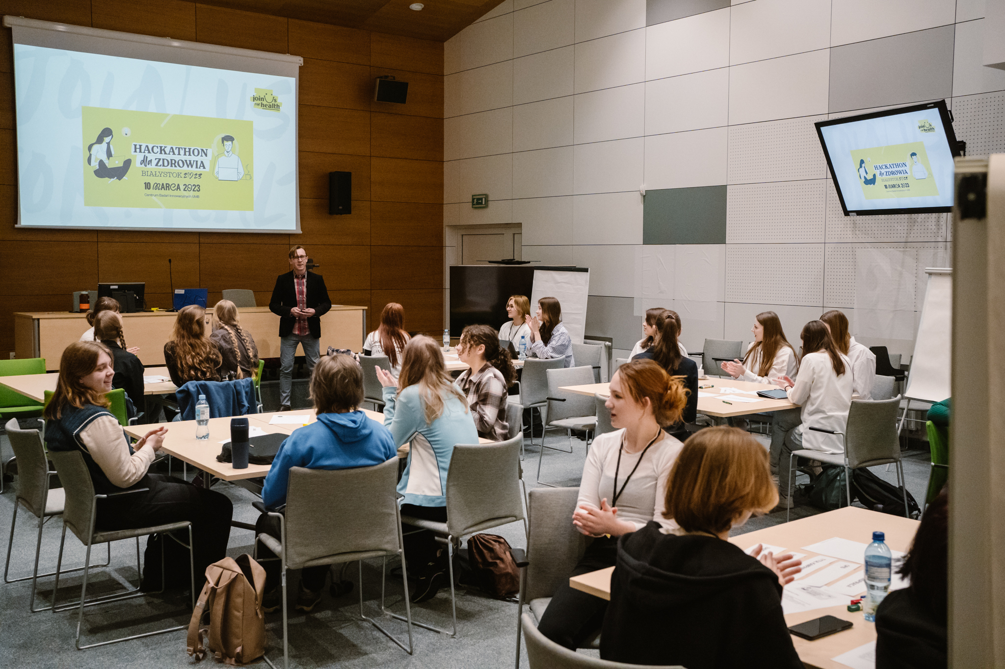 grupa ludzi, uczniów siedzi przy stołach w sali konferencyjnej, w tle ekran