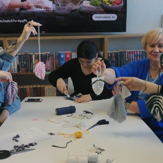 grupa ludzi w różnym wieku siedzi przy stole i robią makramy