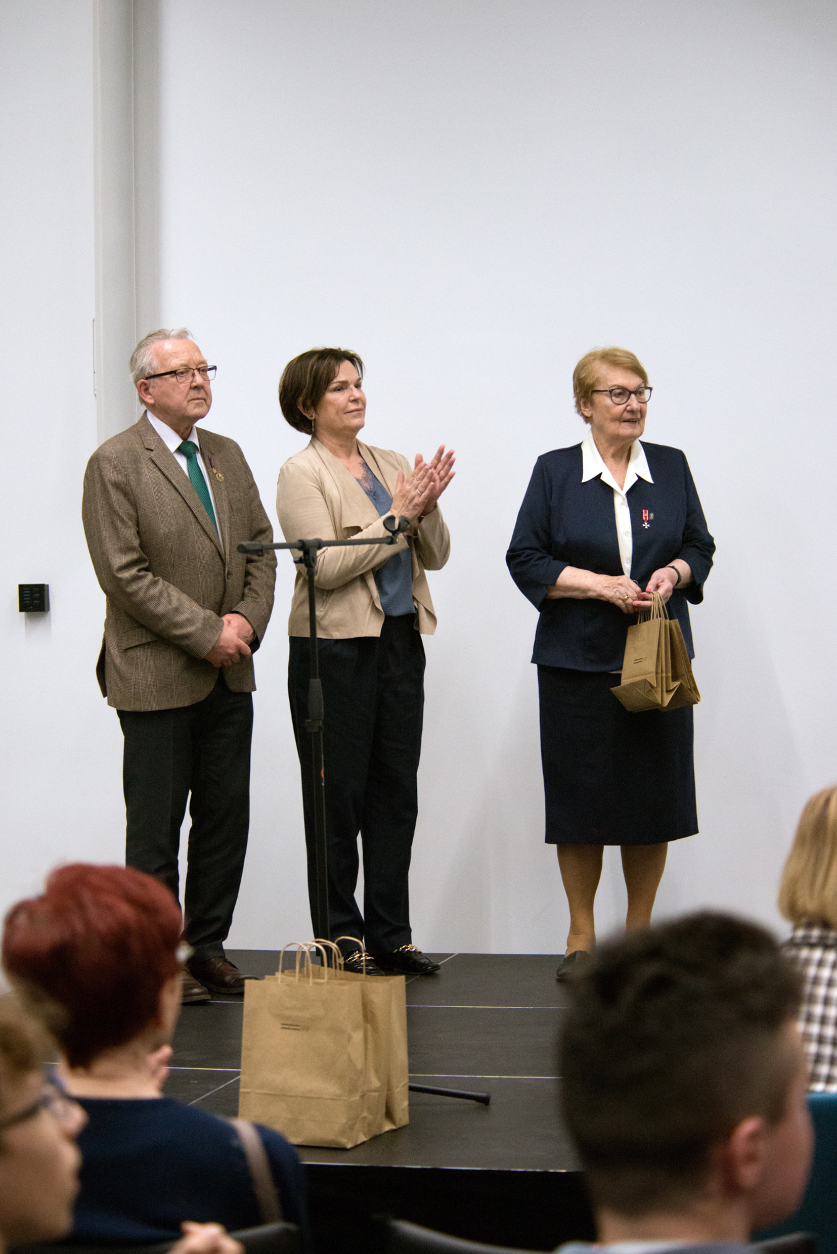 trzy osoby w średnim wieku stoją n scenie, kobieta trzyma torebki papierowe