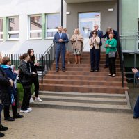 grupa uczniów i nauczycieli stoi na podwórku na placu szkoły, na schodach stoi dyrekcja i zaproszeni goście